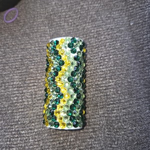 May include: A cylindrical object covered in green and yellow rhinestones. The rhinestones are arranged in a pattern, with a white base. The object is likely a decorative item or accessory.