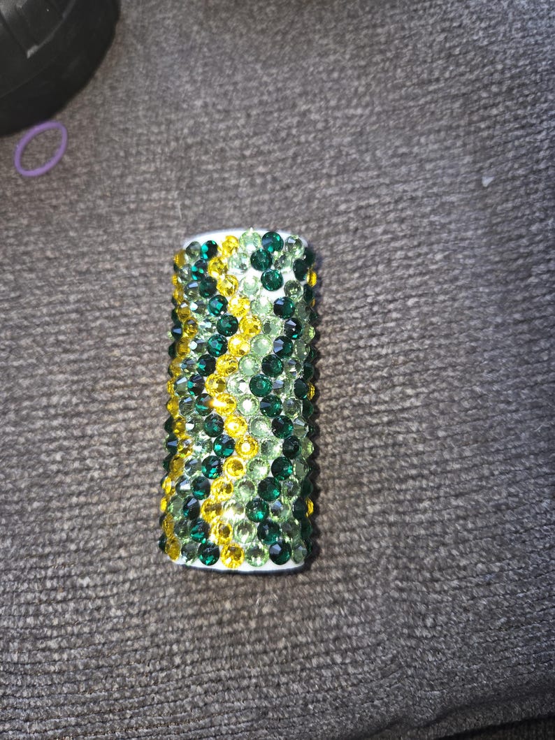 May include: A cylindrical object covered in green and yellow rhinestones. The rhinestones are arranged in a pattern, with a white base. The object is likely a decorative item or accessory.