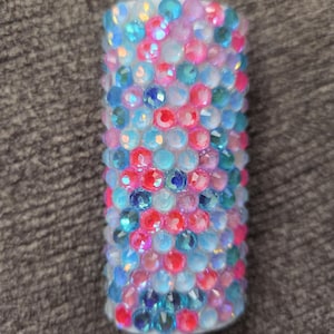 May include: A cylindrical object covered in colorful, sparkling rhinestones. The rhinestones are in shades of blue, pink, and clear, creating a dazzling effect. The object is likely a decorative item or accessory.