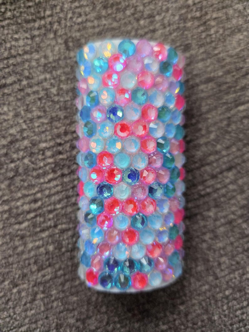 May include: A cylindrical object covered in colorful, sparkling rhinestones. The rhinestones are in shades of blue, pink, and clear, creating a dazzling effect. The object is likely a decorative item or accessory.