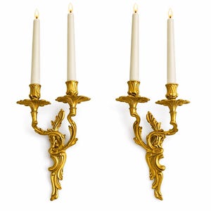 Puede incluir: Dos apliques de pared dorados y ornamentados, cada uno con dos velas encendidas de color marfil. Los apliques presentan un diseño detallado con volutas y detalles florales, creando una estética elegante y clásica. Las velas tienen un brillo suave y cálido.