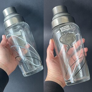 May include: A vintage cocktail shaker with a stainless steel top and a clear glass body. The glass features black and white curved lines and measurement markings. The shaker has a metal label that reads "Cocktail Maison".