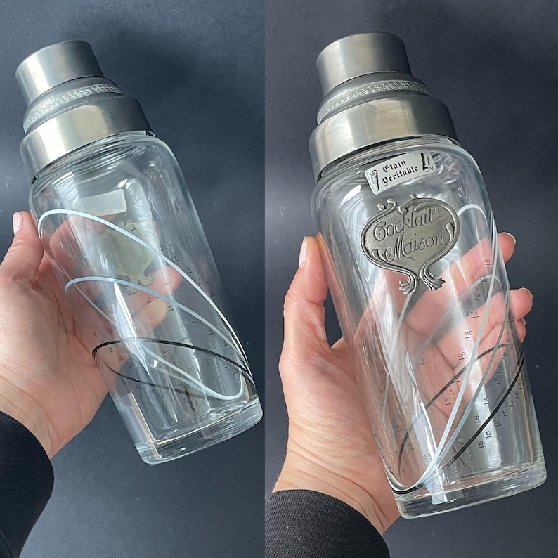 May include: A vintage cocktail shaker with a stainless steel top and a clear glass body. The glass features black and white curved lines and measurement markings. The shaker has a metal label that reads "Cocktail Maison".