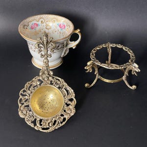 May include: A white porcelain teacup with gold trim and pink rose accents, a brass tea strainer featuring an ornate fleur-de-lis handle and intricate filigree, and a matching brass tripod stand with decorative dragon-like head motifs.