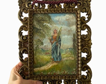 May include: Ornate gold-toned framed religious artwork. The painting depicts a woman in a blue robe holding a palm branch. The frame has an intricate, scrolling design. A burgundy rope is attached for hanging. The artwork is a vintage piece.