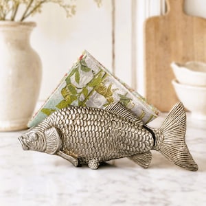 Può includere: Un portatovaglioli in metallo color argento a forma di pesce con squame, pinne e coda dettagliate. Il supporto contiene un tovagliolo di carta piegato con una stampa botanica verde e bianca e piccoli accenti rossi sui bordi della carta.