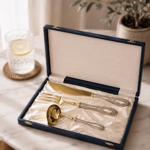 Pode incluir: Um conjunto de três utensílios dourados, incluindo uma faca, um garfo e uma pequena concha, cada um com cabos prateados ornamentados com detalhes em relevo. As peças estão dispostas dentro de uma caixa retangular azul-marinho.