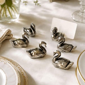 Puede incluir: Un conjunto de cinco tarjeteros de metal plateado con forma de cisne. Cada cisne presenta un cuello curvado y una ranura en la parte posterior diseñada para sostener una pequeña tarjeta blanca rectangular. Las figuras tienen un acabado pulido.