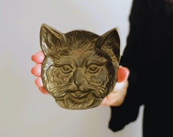 Puede incluir: Un objeto decorativo con forma de gato de color bronce. La cara del gato está detallada con ojos, nariz y boca sonriente. El objeto está sostenido en la mano de una persona, con el fondo desenfocado.