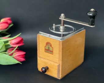 Puede incluir: Un molinillo de café de madera vintage con una manivela de metal y un pequeño cajón. El molinillo tiene una parte superior transparente y un mango negro. La marca "Trusser Mokka" es visible en la parte delantera. Tulipanes rojos en el fondo.