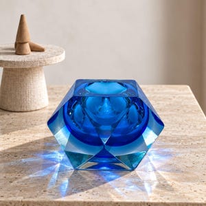 Puede incluir: Un cuenco de cristal azul cobalto facetado con un exterior geométrico de múltiples caras y una abertura interior circular lisa. La construcción de cristal grueso crea refracciones de luz azul profunda y sombras.