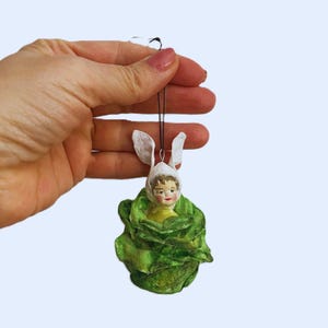 Peut inclure: Ornement de Pâques fait à la main. Il représente une figurine de poupée portant un bonnet blanc avec des oreilles de lapin, nichée dans une structure verte en forme de chou. L'ornement est suspendu par une ficelle noire.