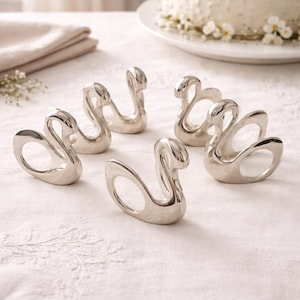 May include: Six silver swan-shaped napkin rings arranged on a white tablecloth. The rings have a smooth, polished finish and are designed with a graceful swan silhouette. A white cake with floral decorations is in the background.
