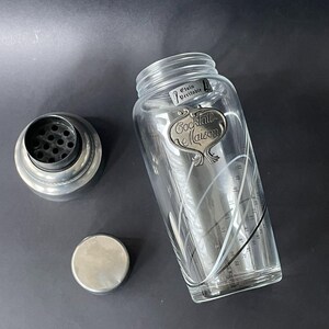 May include: A clear glass cocktail shaker with a silver metal lid, strainer, and base. The shaker has a decorative label that reads "Cocktail Maison" and measurement markings. The words "Etain Veritable" are also visible.