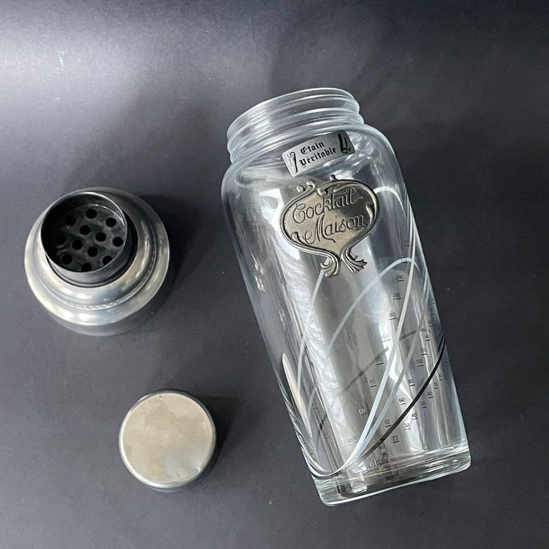 May include: A clear glass cocktail shaker with a silver metal lid, strainer, and base. The shaker has a decorative label that reads "Cocktail Maison" and measurement markings. The words "Etain Veritable" are also visible.