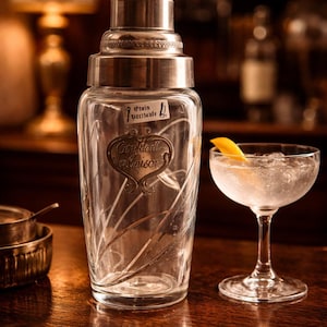 May include: A vintage cocktail shaker with a silver lid and a clear glass body, featuring an etched design and the words "Cocktail Maison." A cocktail glass with ice and a lemon peel sits nearby. The scene is set on a wooden bar.