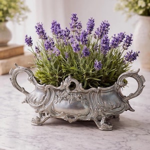 Puede incluir: Una jardinera plateada y ornamentada, llena de lavanda morada vibrante. La jardinera tiene asas decorativas y se apoya en pequeños pies. La lavanda es exuberante y verde, con numerosas espigas de flores moradas. La jardinera está sobre una superficie de mármol.