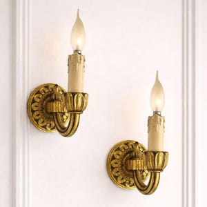 Könnte beinhalten: Zwei verzierte goldene Wandleuchten mit kerzenförmigen Lichtern. Jede Wandleuchte hat eine runde, gemusterte Rückplatte, einen gebogenen Arm und ein kerzenförmiges Licht mit einer mattierten Spitze. Die Wandleuchten sind an einer weißen Wand befestigt.