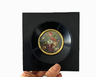 May include: A small, round painting of flowers in a vase, set within a black frame. The painting features red, white, and yellow flowers. The frame is set on a larger square black background.
