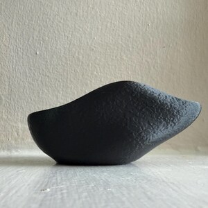 May include: A small, matte black bowl with an organic, asymmetrical shape and a textured, granular surface. The vessel features a curved, elongated rim that rises to a point on one side, creating a distinct, sculptural silhouette.