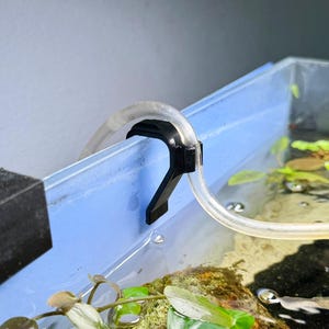 Peut inclure: Un support de tuyau d'aquarium en plastique noir en forme de crochet, conçu pour fixer un tube à air flexible et transparent sur le bord d'une paroi d'aquarium en verre. Le support présente une section supérieure incurvée pour le maintien.