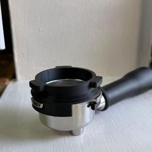 May include: A stainless steel espresso portafilter featuring a black plastic handle and a black circular dosing funnel attachment secured to the top rim. The metal basket is visible inside the portafilter, which has a polished finish.