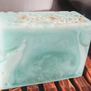 May include: A rectangular bar of soap with a marbled light green and white finish. The top surface of the soap is textured and decorated with small, scattered gold-colored metallic flakes and clear crystalline particles embedded in the surface.