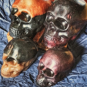 May include: Four decorative skull sculptures in shades of black, orange, and burgundy. The skulls are arranged on a blue textured surface. The skulls have detailed facial features and a glossy finish.