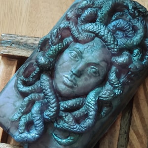 May include: A rectangular bar of soap featuring a raised, three-dimensional relief of a face with snakes for hair. The soap has a dark, iridescent finish with shifting hues of deep purple, teal, and metallic green across the sculpted surface.