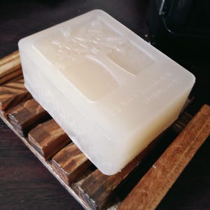 May include: A rectangular, translucent off-white bar of soap featuring an embossed tree design on its top surface. The soap rests on a slatted, dark brown wooden soap dish with visible grain patterns and a smooth, polished finish.