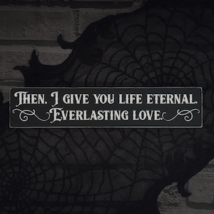 May include: A black wooden sign with white lettering that reads, "Then, I give you life eternal. Everlasting love." The sign has decorative flourishes and is set against a dark, textured background.