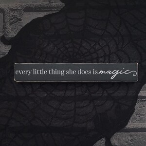 May include: A rectangular, black wooden sign with white text that reads "every little thing she does is magic". The sign is set against a dark, textured background, with a brick-like pattern visible on the left.