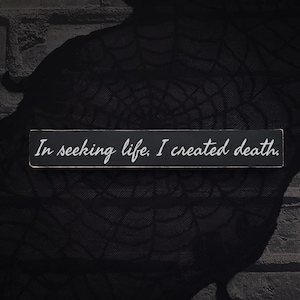 May include: A rectangular black wooden sign with the text "In seeking life, I created death" in white script. The sign is set against a dark, textured background, creating a stark contrast.