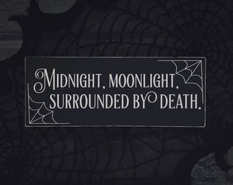 Może przedstawiać: Prostokątny czarny drewniany znak z białym tekstem: „MIDNIGHT, MOONLIGHT, SURROUNDED BY DEATH”. Znak ma dekoracyjne pajęczyny w rogach. Tło to ciemna, teksturowana tkanina.