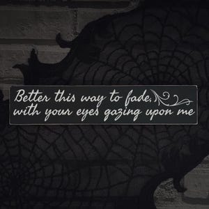 May include: A rectangular black wooden sign with white cursive text that reads "Better this way to fade, with your eyes gazing upon me." The sign is set against a dark background with a spiderweb design.