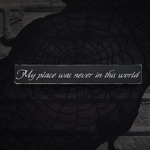May include: A rectangular black wooden sign with the text "My place was never in this world" in white cursive lettering. The sign is set against a dark, textured background.