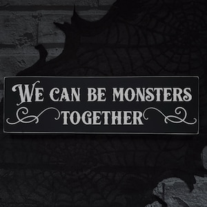 May include: A rectangular black sign with white text that reads "We Can Be Monsters Together." The sign has decorative flourishes and is likely for Halloween or a similar theme. The background is dark.