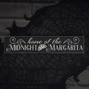 以下が含まれることがあります： 「home of the MIDNIGHT MARGARITA」と白い文字で書かれた長方形の黒い木製サイン。装飾的な装飾とライムのスライスグラフィックが特徴です。暗い背景に表示されています。