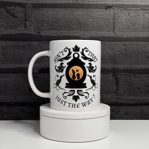 May include: White ceramic mug with a black and yellow design. The design features a silhouette of two figures inside a lantern, with the text "Ain't that just the way?" in a decorative font. The mug has a curved handle.