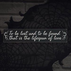 May include: A rectangular black wooden sign with white text that reads, "To be lost and to be found that is the lifespan of love." The sign has decorative white accents on each end. The background is a dark, textured fabric.