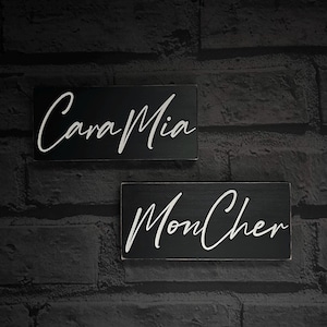 May include: Two rectangular black wooden signs with white cursive text. The top sign reads "Cara Mia", and the bottom sign reads "Mon Cher". The signs are mounted on a dark gray brick wall.