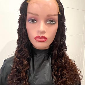 May include: A mannequin head with a dark brown, curly wig. The wig features a lace front and is styled with loose, voluminous waves. The mannequin has painted features, including red lips and eye makeup. The mannequin is wearing a black cape.