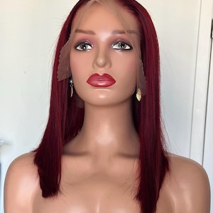 May include: A burgundy-colored wig with a lace front is displayed on a mannequin head. The wig features a straight, shoulder-length style. The mannequin has painted features, including red lipstick and eye makeup. The wig is a deep, rich shade of red.
