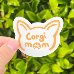 May include: White sticker with an orange outline of a dog's head. The text "Corgi mom" is written in orange inside the outline.