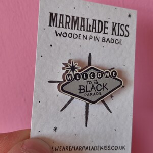 May include: A wooden pin badge with the text "WELCOME to The BLACK PARADE" in a gray and black design. The pin is in the shape of a sign with a starburst design. The pin is on a white card with the text "MARMALADE KISS WOODEN PIN BADGE".