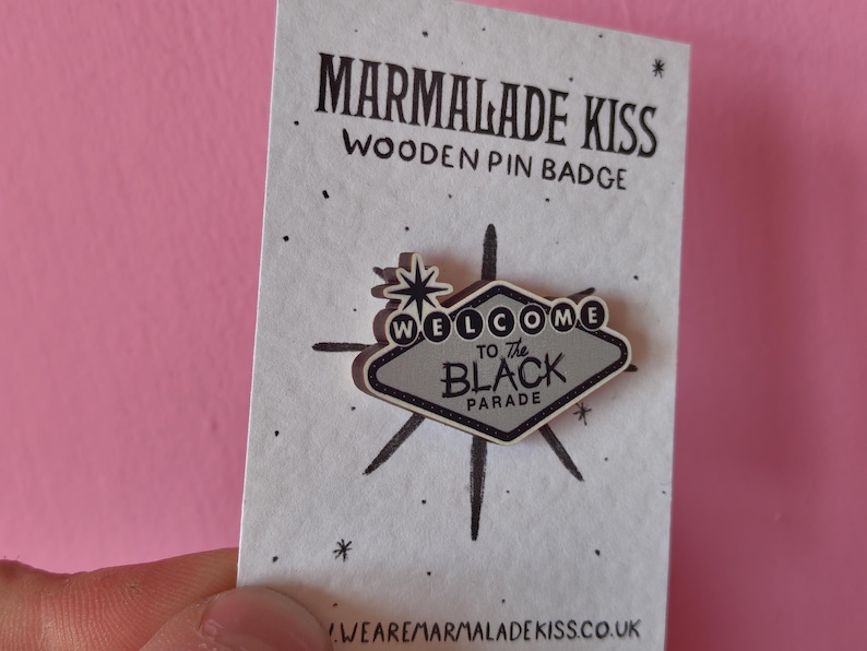 May include: A wooden pin badge with the text "WELCOME to The BLACK PARADE" in a gray and black design. The pin is in the shape of a sign with a starburst design. The pin is on a white card with the text "MARMALADE KISS WOODEN PIN BADGE".