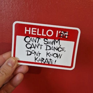May include: Red and white name tag with the text "HELLO I'M CAN'T SWIM, CAN'T DANCE, DON'T KNOW KARATE!"