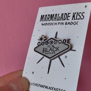 May include: Wooden pin badge with a retro design. The pin features a gray sign with the words "WELCOME TO THE BLACK PARADE" and a starburst design. The pin is on a white card with the words "MARMALADE KISS" printed in black.