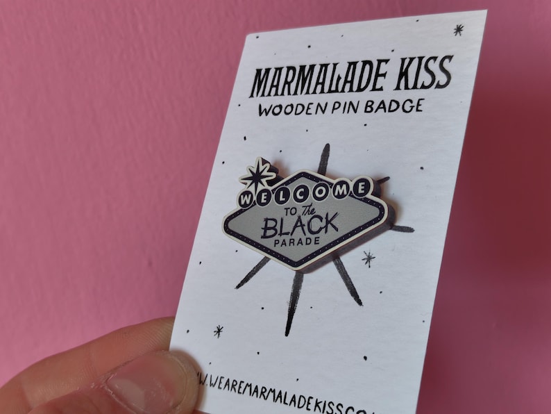 May include: Wooden pin badge with a retro design. The pin features a gray sign with the words "WELCOME TO THE BLACK PARADE" and a starburst design. The pin is on a white card with the words "MARMALADE KISS" printed in black.