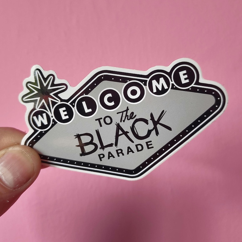 K&ouml;nnte beinhalten: Ein Aufkleber mit einem schwarz-wei&szlig;en Design mit der Aufschrift "WELCOME to The BLACK PARADE". Der Aufkleber hat die Form eines Retro-Schildes mit einem Stern auf der linken Seite. Der Hintergrund ist rosa.